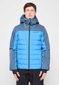 Mann trägt eine zweifarbige blaue isolierte Jacke mit Kapuze und schwarze Hose, steht vor einem einfarbigen hellgrauen Hintergrund.