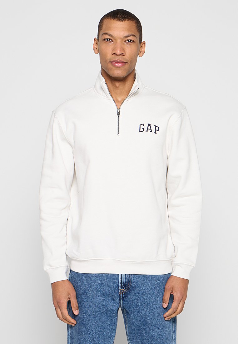 Weißes Sweatshirt mit Reißverschluss am Kragen und navyfarbenem "GAP"-Logo auf der Brust, aus weichem Stoff, mit gerippten Bündchen und Saum, getragen zu blauen Jeans.