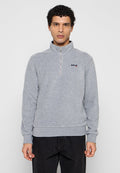 Mann mit kurzen lockigen Haaren, der einen grauen Pullover mit Viertel-Reißverschluss und Schott NYC-Logo sowie schwarze Hosen trägt, steht vor einem schlichten hellen Hintergrund.