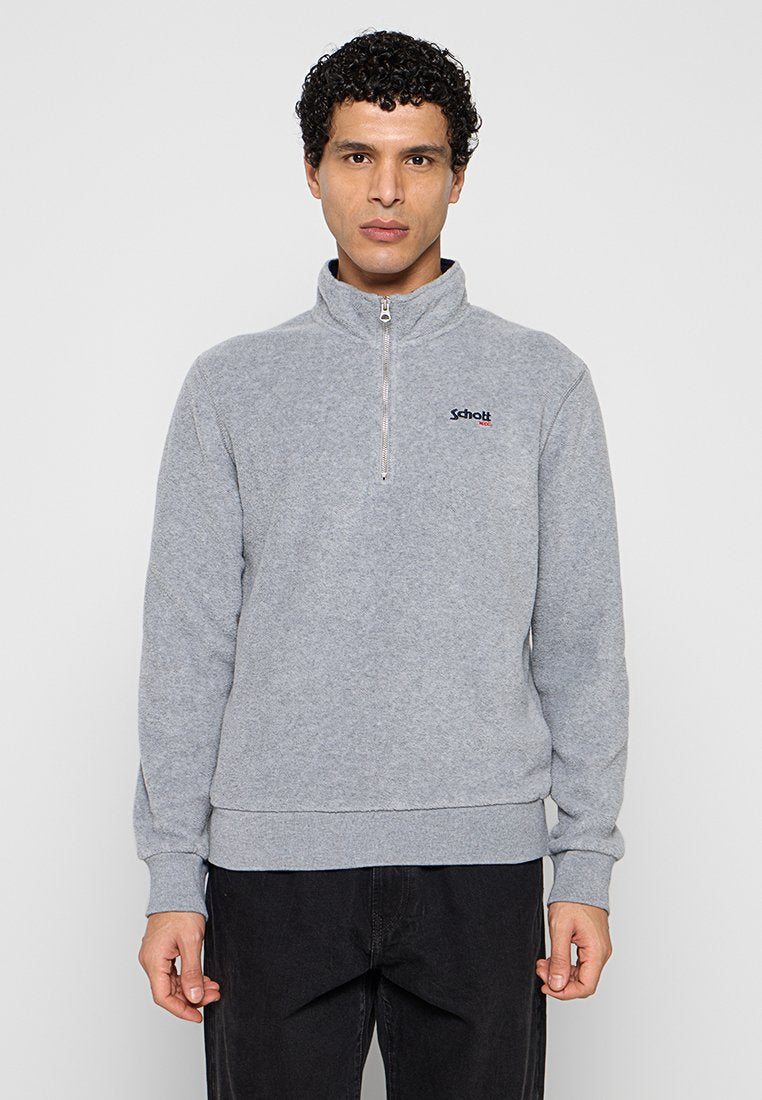 Mann mit kurzen lockigen Haaren, der einen grauen Pullover mit Viertel-Reißverschluss und Schott NYC-Logo sowie schwarze Hosen trägt, steht vor einem schlichten hellen Hintergrund.