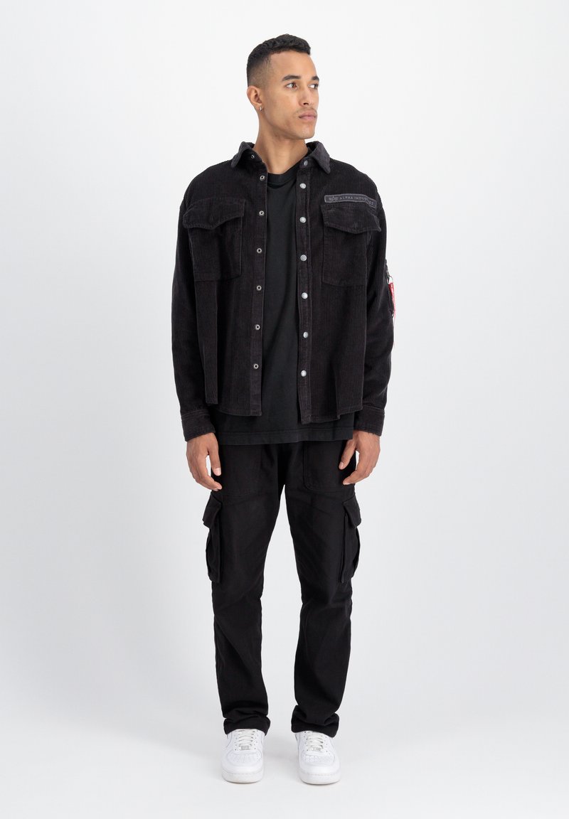 Schwarze Cordjacke mit Vordertaschen, über einem schwarzen Hemd und schwarzen Cargo-Hosen getragen. Weiße Sneakers vervollständigen das Outfit.