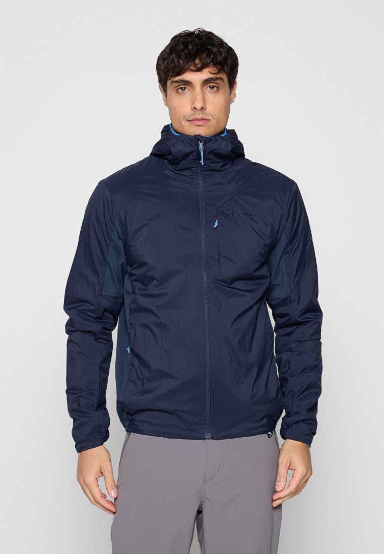 Mann trägt eine marineblaue Kapuzenjacke mit Reißverschluss und hellgraue Hose, steht frontal vor einem schlichten Hintergrund.