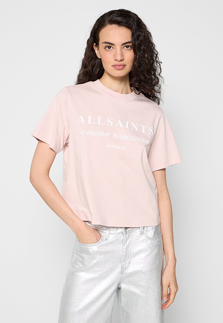 Frau mit lockigem Haar, die ein hellrosa AllSaints T-Shirt und glänzende silberne Hose trägt, posiert mit einer Hand in der Tasche.