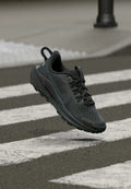 Schwarzer Sportschuh mit Mesh-Obermaterial und Gummisohle, versehen mit strukturierten Akzenten und Schnürsenkeln, in der Luft über einem Zebrastreifen dargestellt.