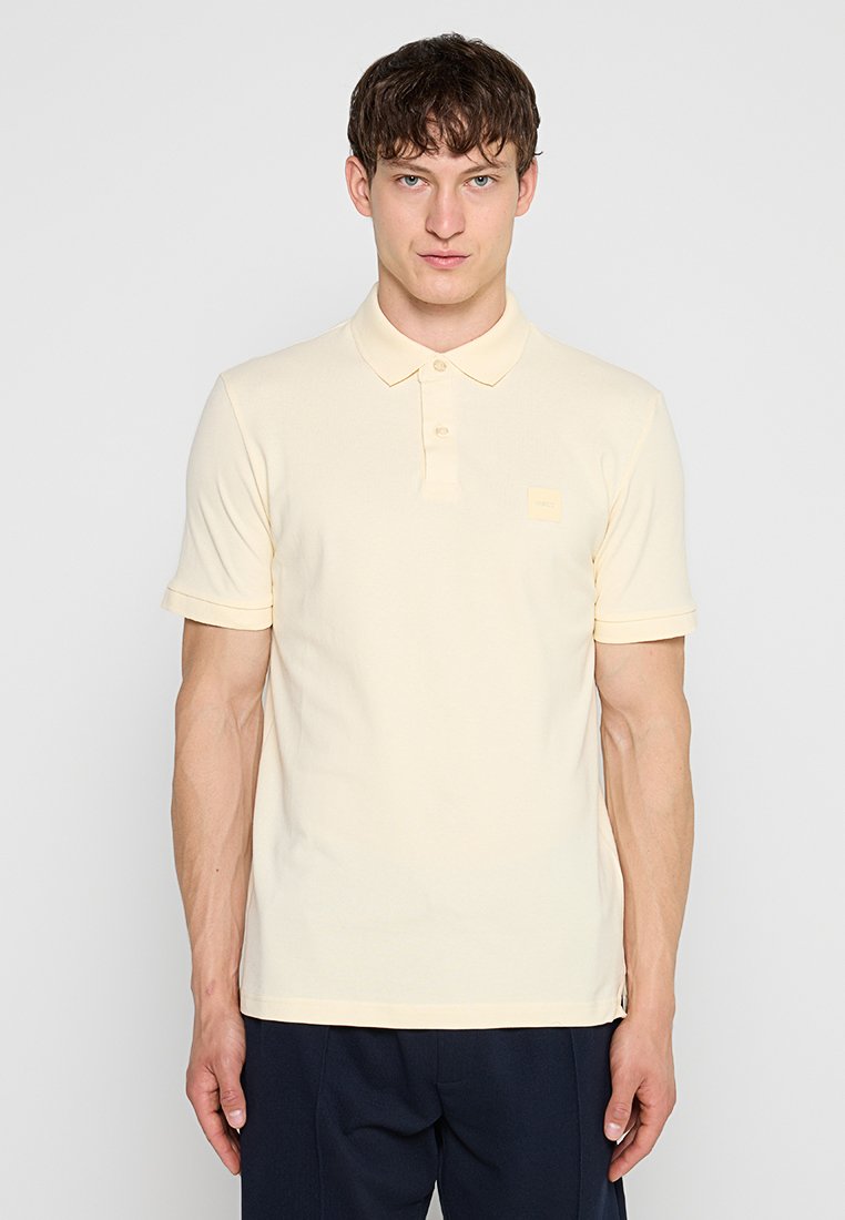 Junger Mann mit kurzen braunen Haaren, der ein hellgelbes kurzärmeliges Poloshirt und dunkle Hosen trägt, steht vor einem einfarbigen grauen Hintergrund.