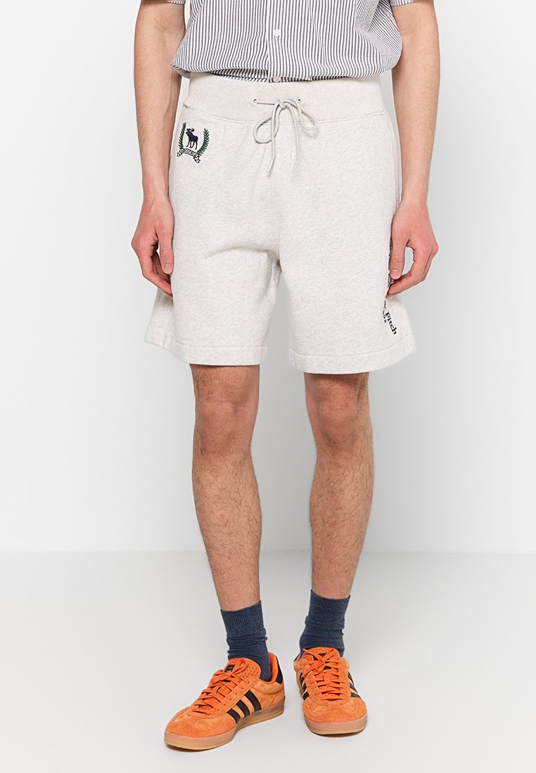 Mann trägt helle graue Shorts mit Kordelzug und besticktem Logo, navyfarbene Socken und leuchtend orangefarbene Sneaker mit schwarzen Streifen, steht vor weißem Hintergrund.