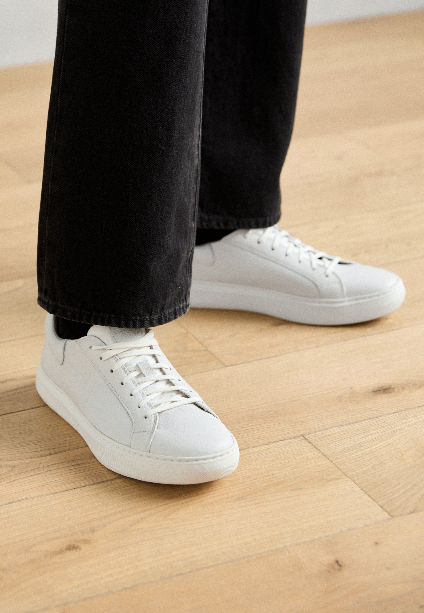 Weißlederen Low-Top-Sneaker mit Schnürung, abgerundeter Zehenpartie und minimalistischem Design. Kombiniert mit schwarzen, schmal zulaufenden Jeans.