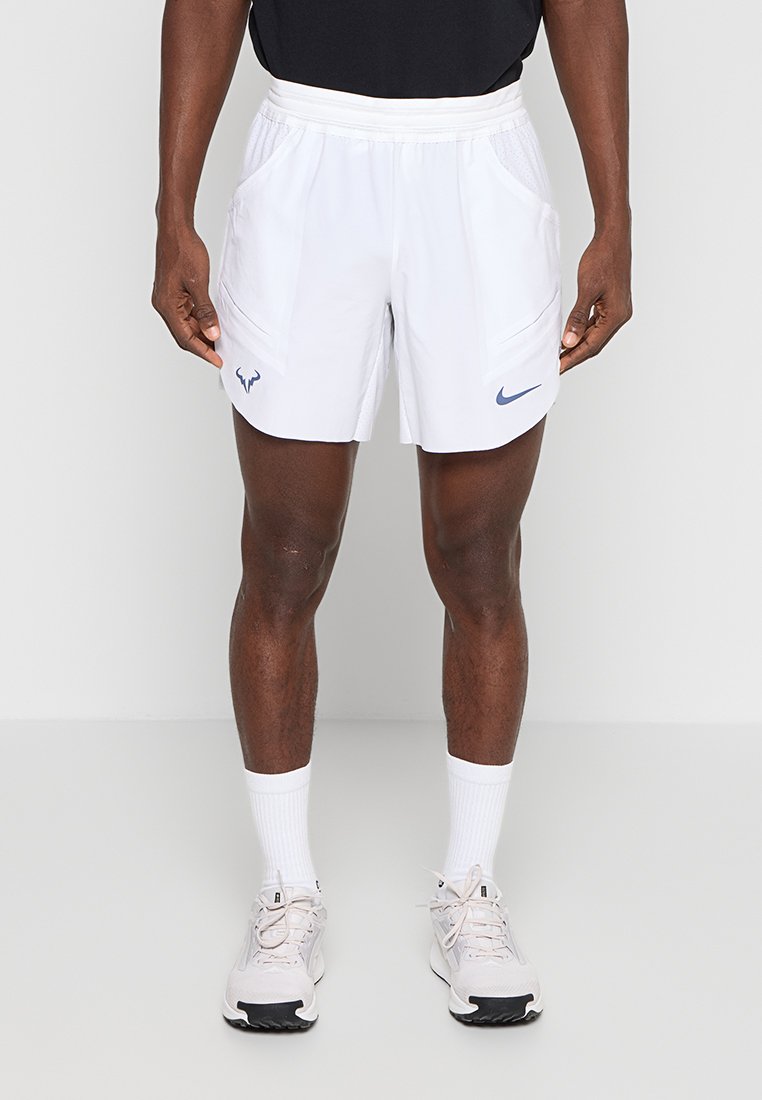 Athlet trägt weiße Nike-Sportshorts mit Logos, weiße Socken und weiße Laufschuhe, steht vor einem schlichten hellen Hintergrund.