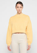 Gelbes Cropped-Sweatshirt mit langen Ärmeln und hohem Kragen. Weiche Textur mit gerippten Bündchen. Kombiniert mit hellbeigen Leggings.