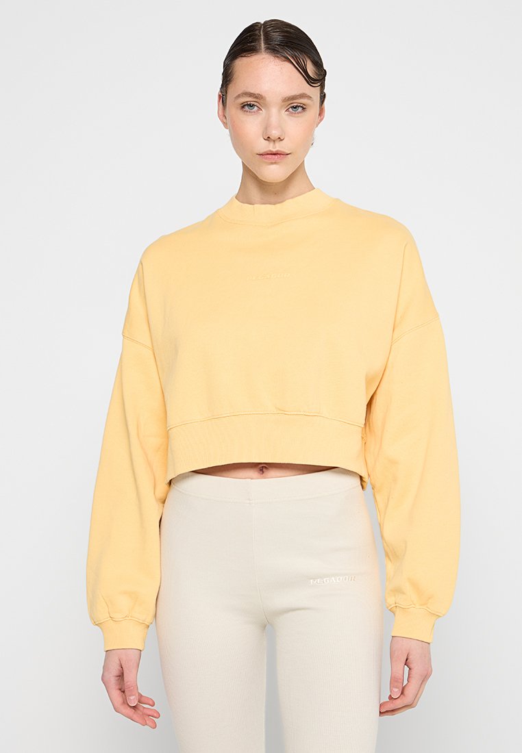 Gelbes Cropped-Sweatshirt mit langen Ärmeln und hohem Kragen. Weiche Textur mit gerippten Bündchen. Kombiniert mit hellbeigen Leggings.