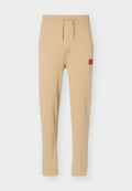 Beige zulaufende Jogginghose mit elastischem Bund, Kordelzug, Seitentaschen, gerippten Bündchen am Knöchel und rotem Logopatch am rechten Oberschenkel.