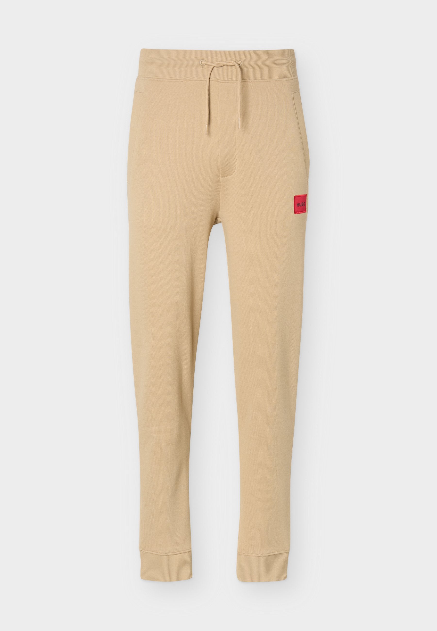 Beige zulaufende Jogginghose mit elastischem Bund, Kordelzug, Seitentaschen, gerippten Bündchen am Knöchel und rotem Logopatch am rechten Oberschenkel.