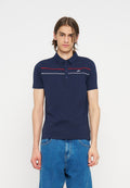 Junger Mann mit braunen Haaren, der ein dunkelblaues Poloshirt mit roten und weißen Streifen und blaue Jeans trägt, steht vor einem einfarbigen weißen Hintergrund.