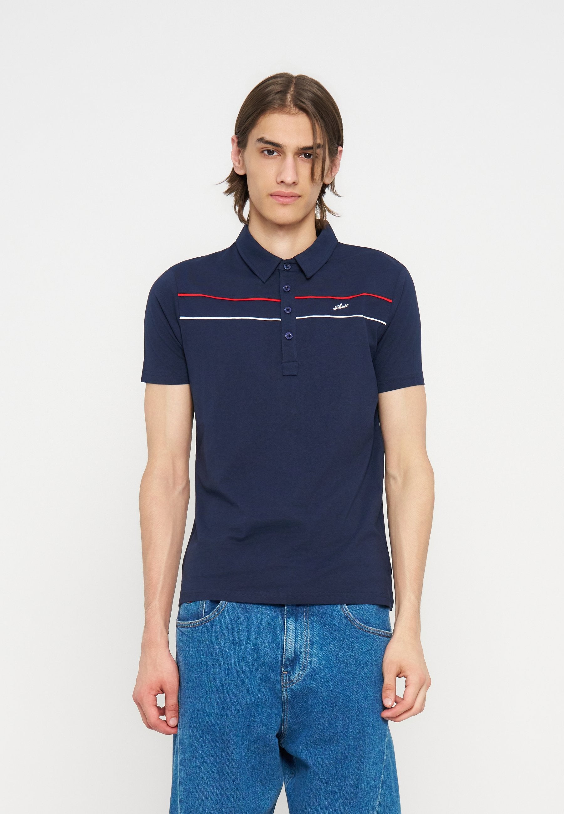 Junger Mann mit braunen Haaren, der ein dunkelblaues Poloshirt mit roten und weißen Streifen und blaue Jeans trägt, steht vor einem einfarbigen weißen Hintergrund.