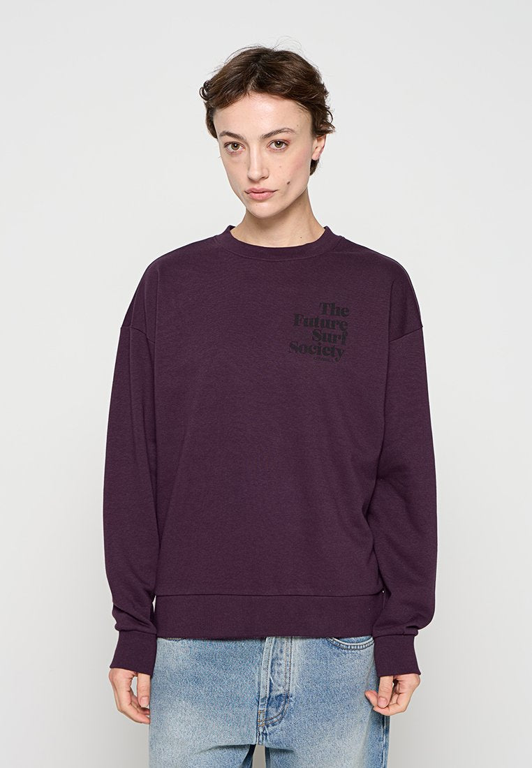 Frau trägt einen dunkelvioletten Sweatshirt mit dem Schriftzug "The Future Surf Society" und hellblaue Jeans, steht vor einem schlichten hellen Hintergrund.