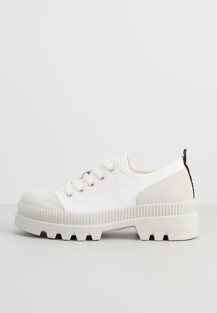 Niedrig geschnittene weiße Sneaker mit dickem, geripptem Absatz, Schnürung vorne und Wildlederakzenten an Zehen und Ferse, seitlich auf einem einfarbigen Hintergrund gezeigt.