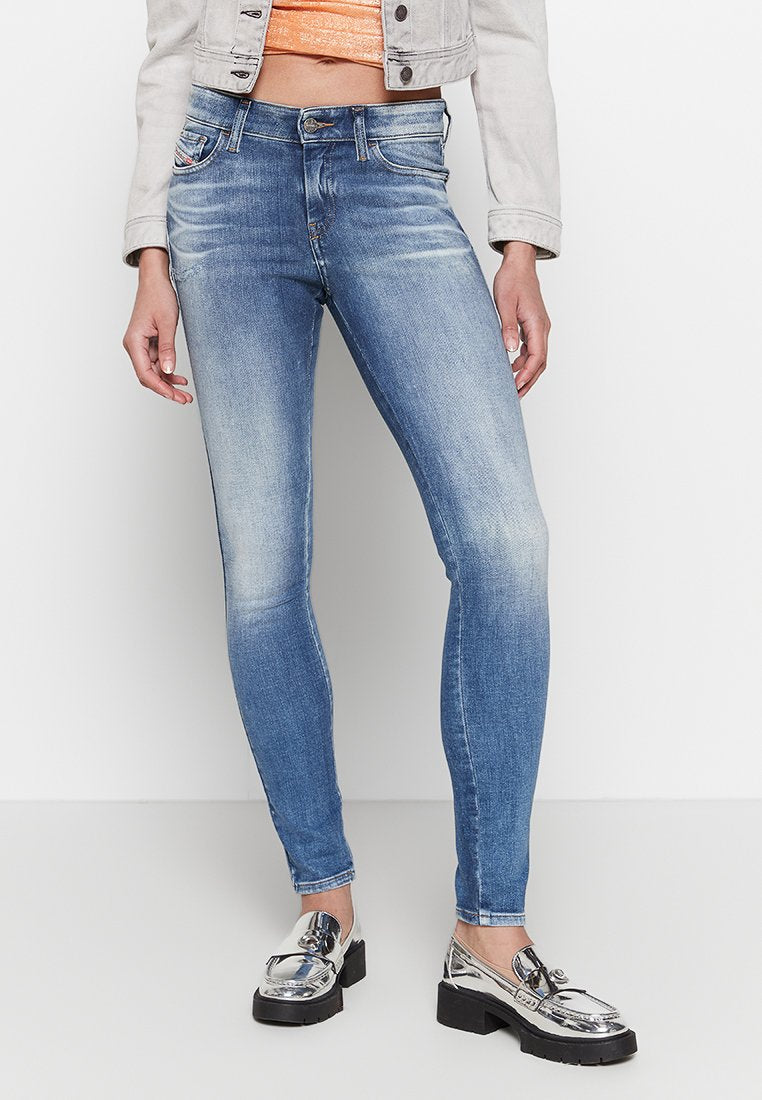 Frau in hellblauen Skinny Jeans, silbernen metallischen Slippers, einer beigefarbenen Jacke in Knielänge und einem orangefarbenen Crop-Top.