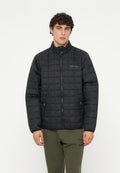 Junger Mann mit lockigem Haar, der eine schwarze gesteppte Reißverschlussjacke und olivgrüne Hose trägt und vor einem schlichten weißen Hintergrund steht.