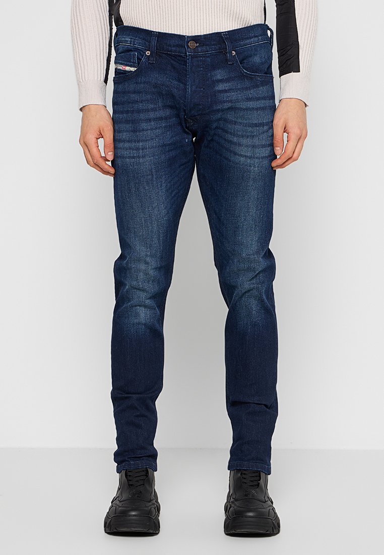 Dunkelblaue Denim-Jeans mit schlanker Passform, sichtbaren Nähten, Vordertaschen und dezenten Verblassungen. Kombiniert mit schwarzen Schuhen.