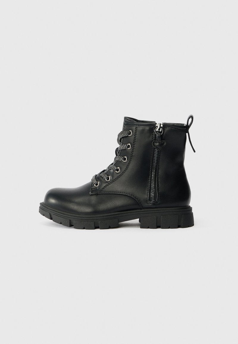 Schwarze Leder-Stiefelette mit dickem Profilsohle, seitlichem Reißverschluss mit Herz-Anhänger und schwarz-weißen Schnürsenkeln vor hellgrauem Hintergrund.