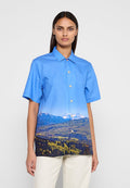 Blaues Kurzarmhemd mit Landschaftsaufdruck von Bergen und Grünflächen. Es hat einen Kragen und eine Knopfleiste vorne, gefertigt aus glattem Stoff.