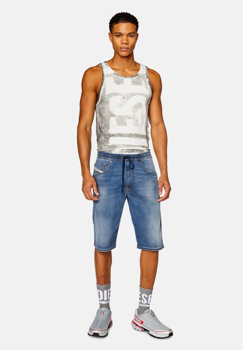 Graues Tanktop mit großem weißen Logo, blaue Jeansshorts und graue Slipper mit roten Akzenten. Das Modell trägt grau-weiße Socken.