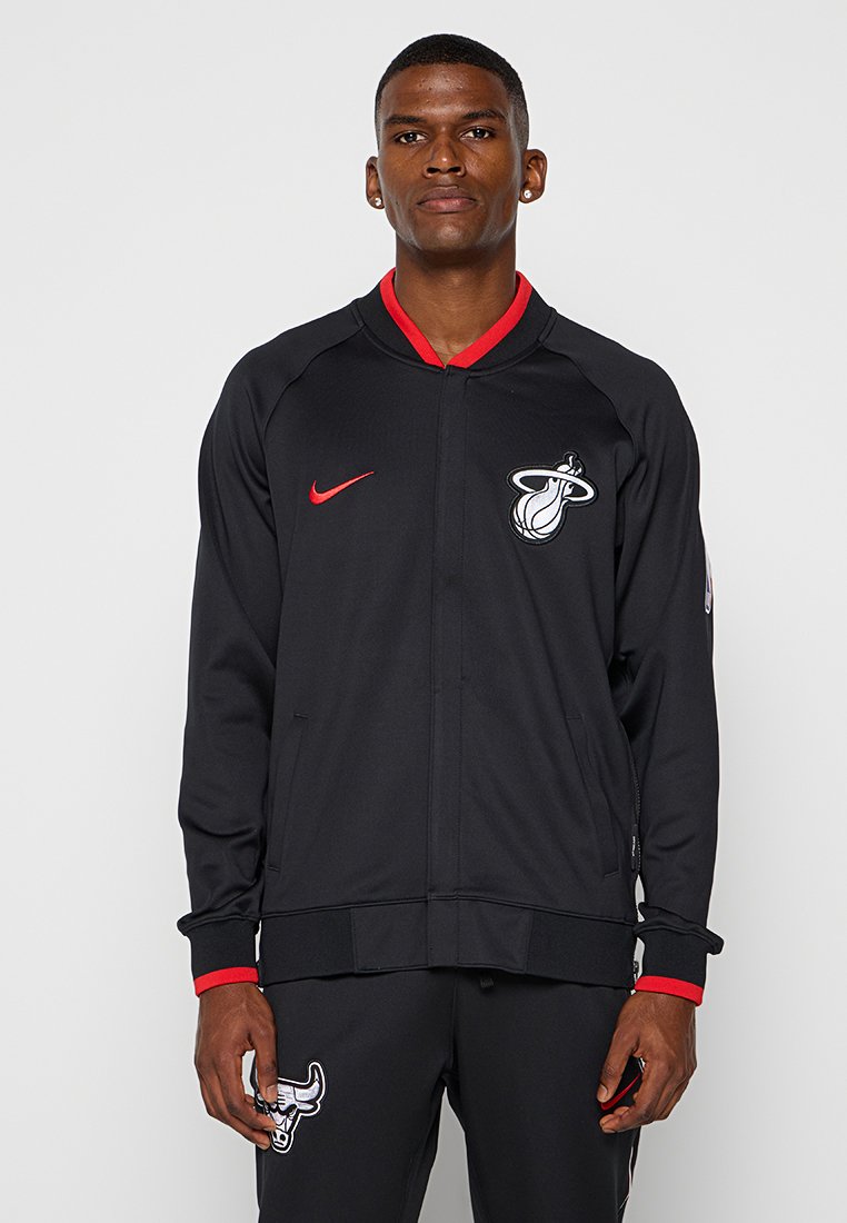 Schwarze Sportjacke mit roten Akzenten, ausgestattet mit einem Reißverschluss vorne, gerippten Bündchen und einem Logo auf der linken Brust. Glattes, leichtes Material.