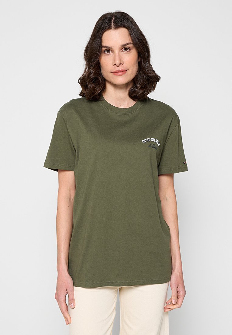 Frau mit schulterlangem braunem Haar, die ein olivgrünes T-Shirt mit kurzen Ärmeln und weißem Text auf der Brust sowie hellbeige Hosen trägt.
