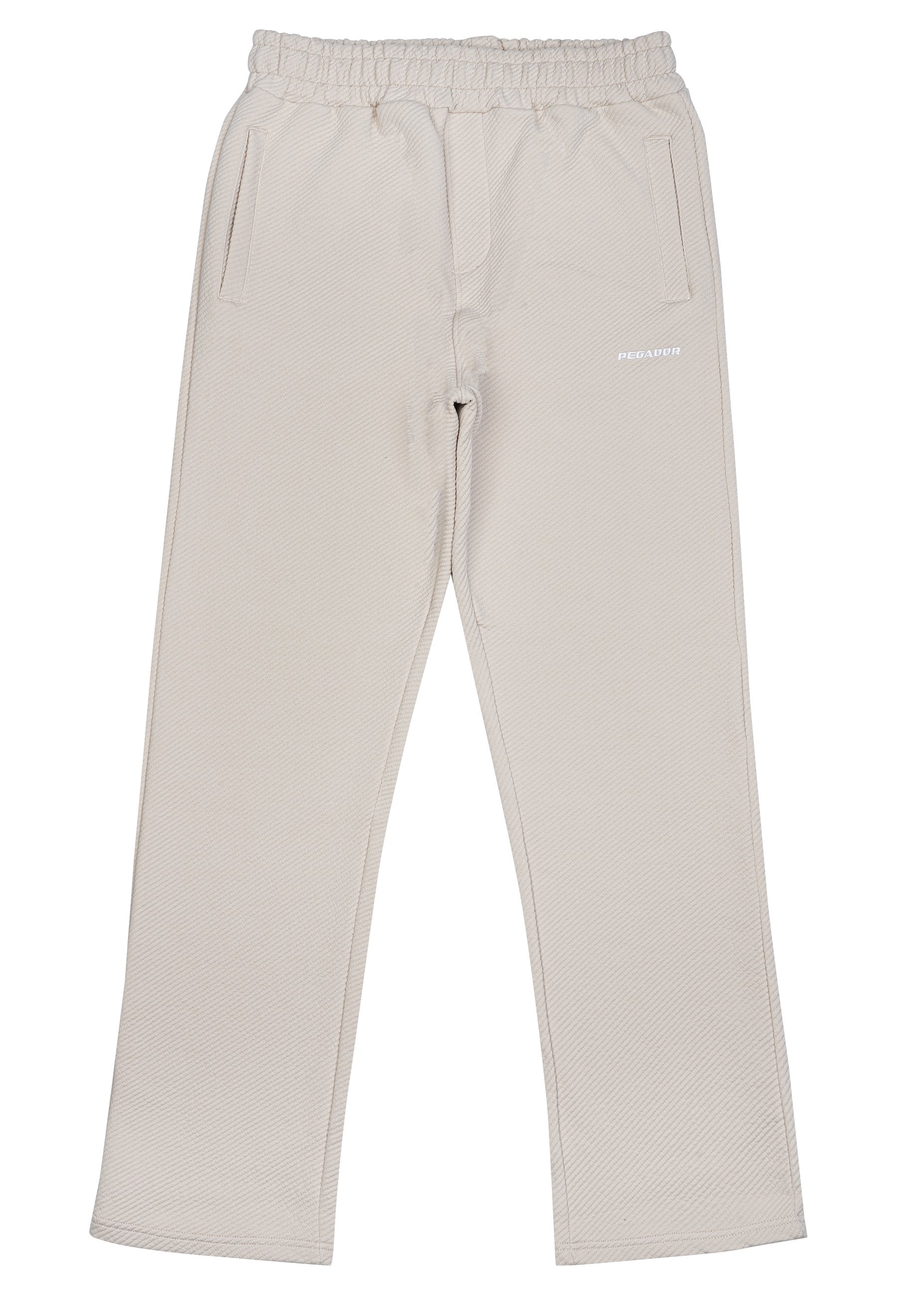 Beige Hose aus strukturiertem Stoff, mit einem elastischen Bund, zwei Seitentaschen und einem kleinen weißen Logo auf der Vorderseite.
