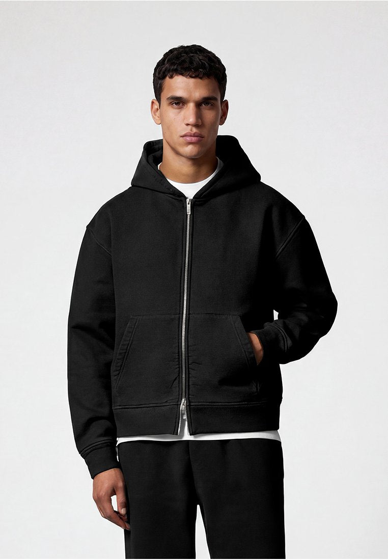 Mann trägt einen schwarzen Kapuzenpullover mit Reißverschluss über einem weißen Hemd, mit einer Hand in der vorderen Tasche, steht vor einem einfarbigen hellen Hintergrund.