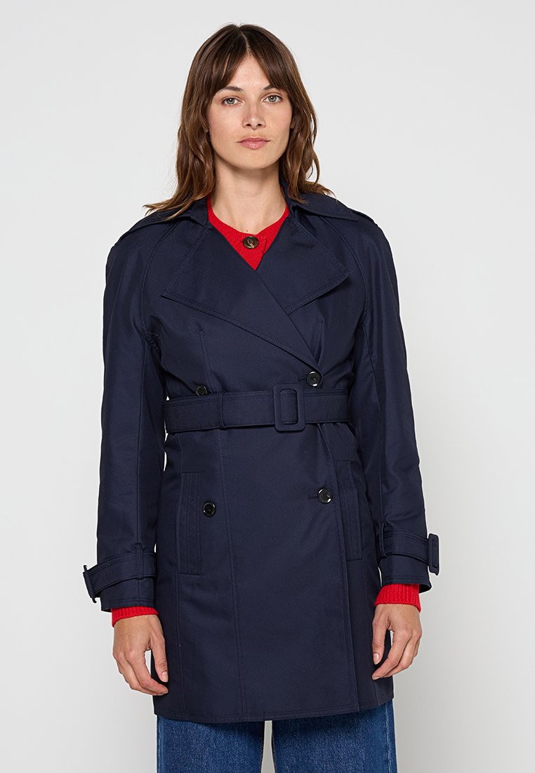 Marinefarbener Trenchcoat mit doppelter Knopfleiste, taillierter Taille, großen Taschen und spitzem Kragen; der Stoff wirkt glatt und wasserabweisend.