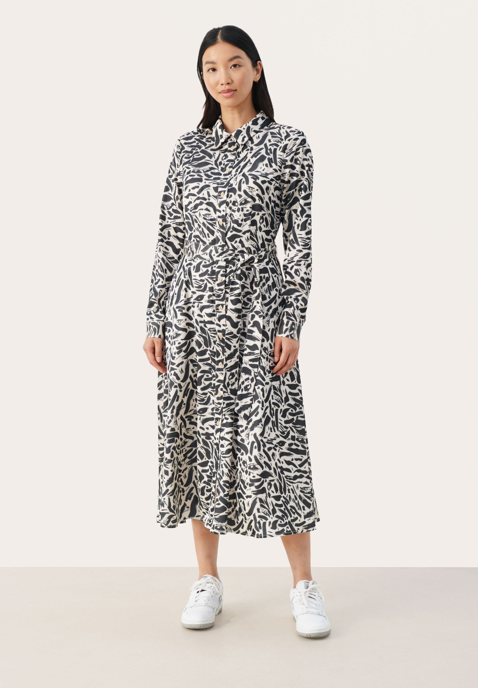 Langärmeliges, knöpfbares Kleid im schwarz-weißen abstrakten Muster; tailliertes Design; leichtes Material mit fließendem Rock; kombiniert mit weißen Sneakers.