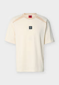 Cremefarbenes T-Shirt mit kurzen Ärmeln, beigen Schulterakzenten und einem kleinen schwarzen quadratischen Logo-Aufnäher mit der Aufschrift "DR HUGO" mittig auf der Brust.