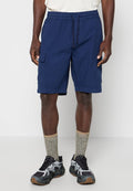 Mann trägt dunkelblaue Cargo-Shorts, graue gemusterte Kniestrümpfe, schwarze und graue Sportschuhe, steht vor einfachem Hintergrund.