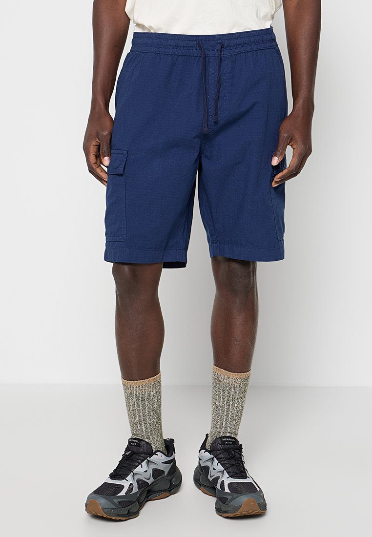 Mann trägt dunkelblaue Cargo-Shorts, graue gemusterte Kniestrümpfe, schwarze und graue Sportschuhe, steht vor einfachem Hintergrund.