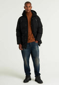 Schwarze gepolsterte Jacke mit Kapuze, Reißverschluss und Druckknopfverschlüssen, zwei Fronttaschen. Kombiniert mit einem braunen Rollkragenpullover und blauen Jeans, abgerundet mit schwarzen Stiefeln.