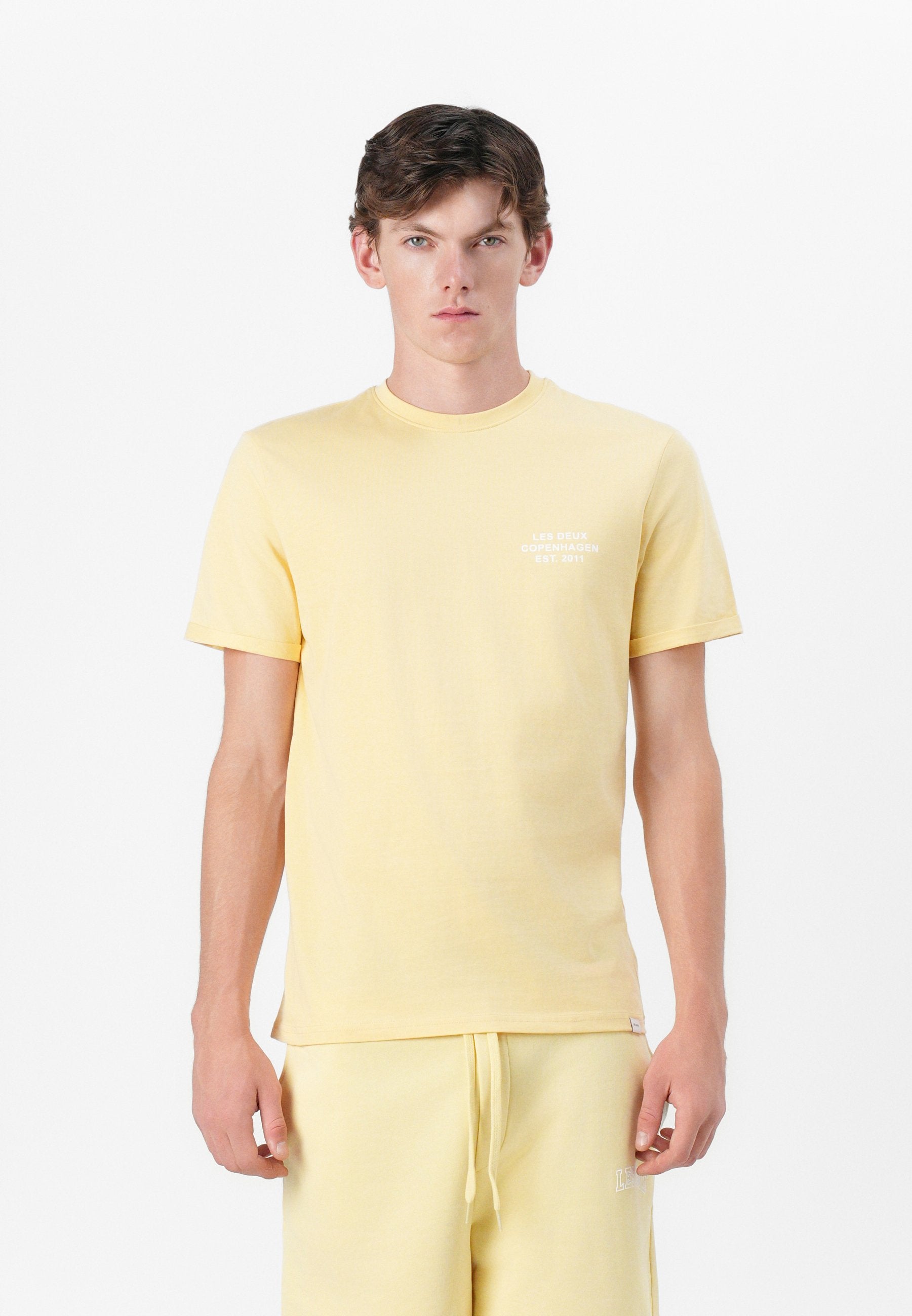 Gelbes Baumwoll-T-Shirt mit Rundhalsausschnitt, kurzen Ärmeln und einem kleinen weißen Logo auf der linken Brust. Weiche Textur, lässige Passform.