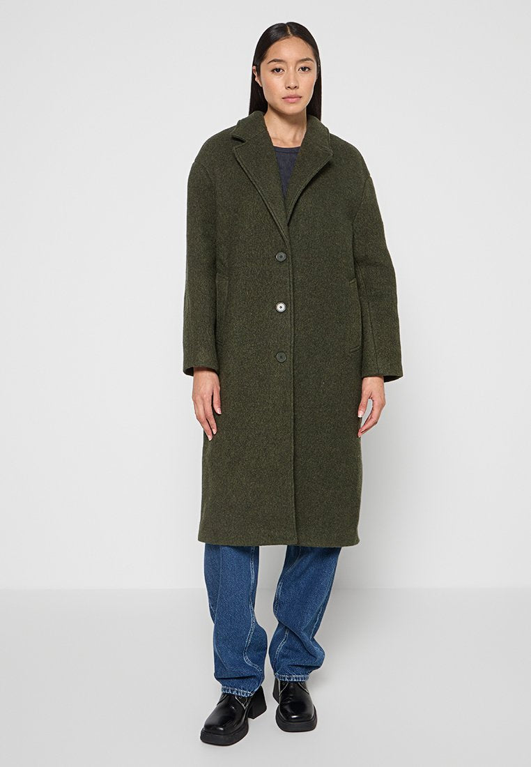 Olivengrüner Wollmix-Mantel mit Reverskragen, vorderen Knöpfen und zwei Seitentaschen, kombiniert mit lässigen Denim und schwarzen Schuhen.