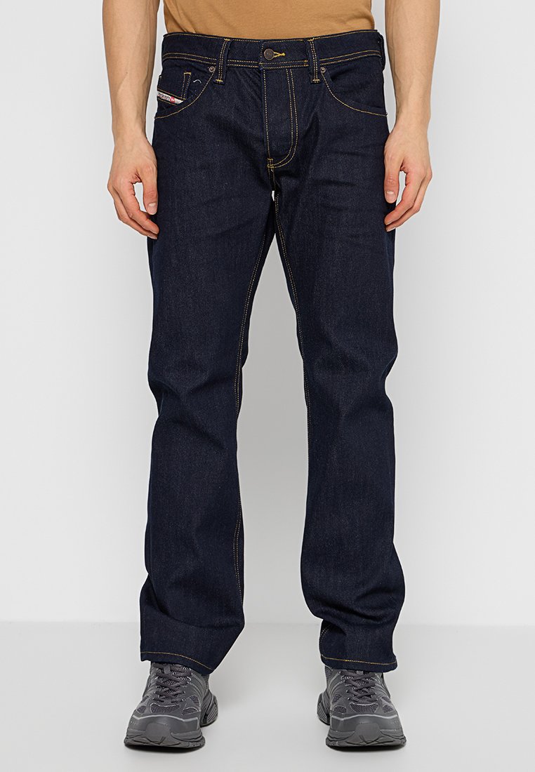 Mann in dunkelblauen, gerade geschnittenen Jeans mit kontrastierenden Nähten und grauen Sportschuhen, der vor einem weißen Hintergrund steht.