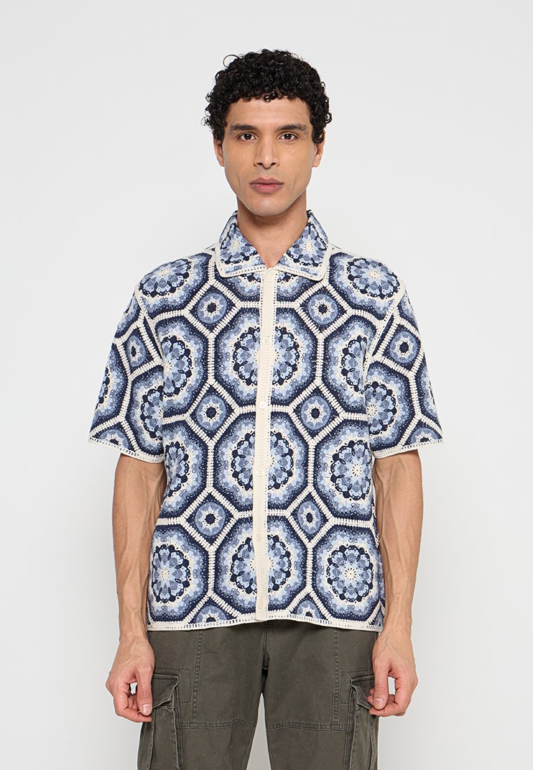 Mann trägt ein kurzärmeliges Hemd mit blauen und weißen geometrischen Blumenmustern und olivgrüne Cargohosen, steht vor einem schlichten Hintergrund.