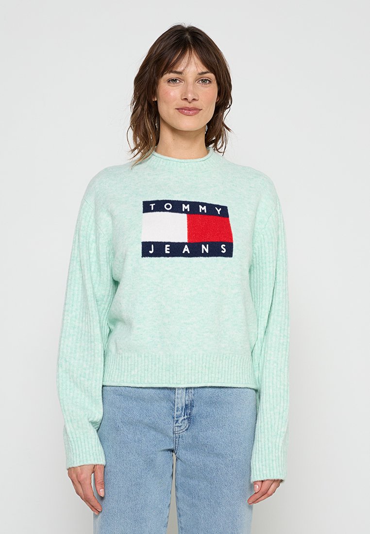 Frau trägt einen mintgrünen Tommy Jeans Pullover mit großem Logo und hellblauer Jeans, steht vor einem schlichten weißen Hintergrund.