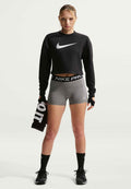 Frau in schwarzem, kurz geschnittenem Sweatshirt und grauen Nike Pro Shorts hält ein schwarzes Handtuch, trägt schwarze Handschuhe, Socken und Laufschuhe und steht.