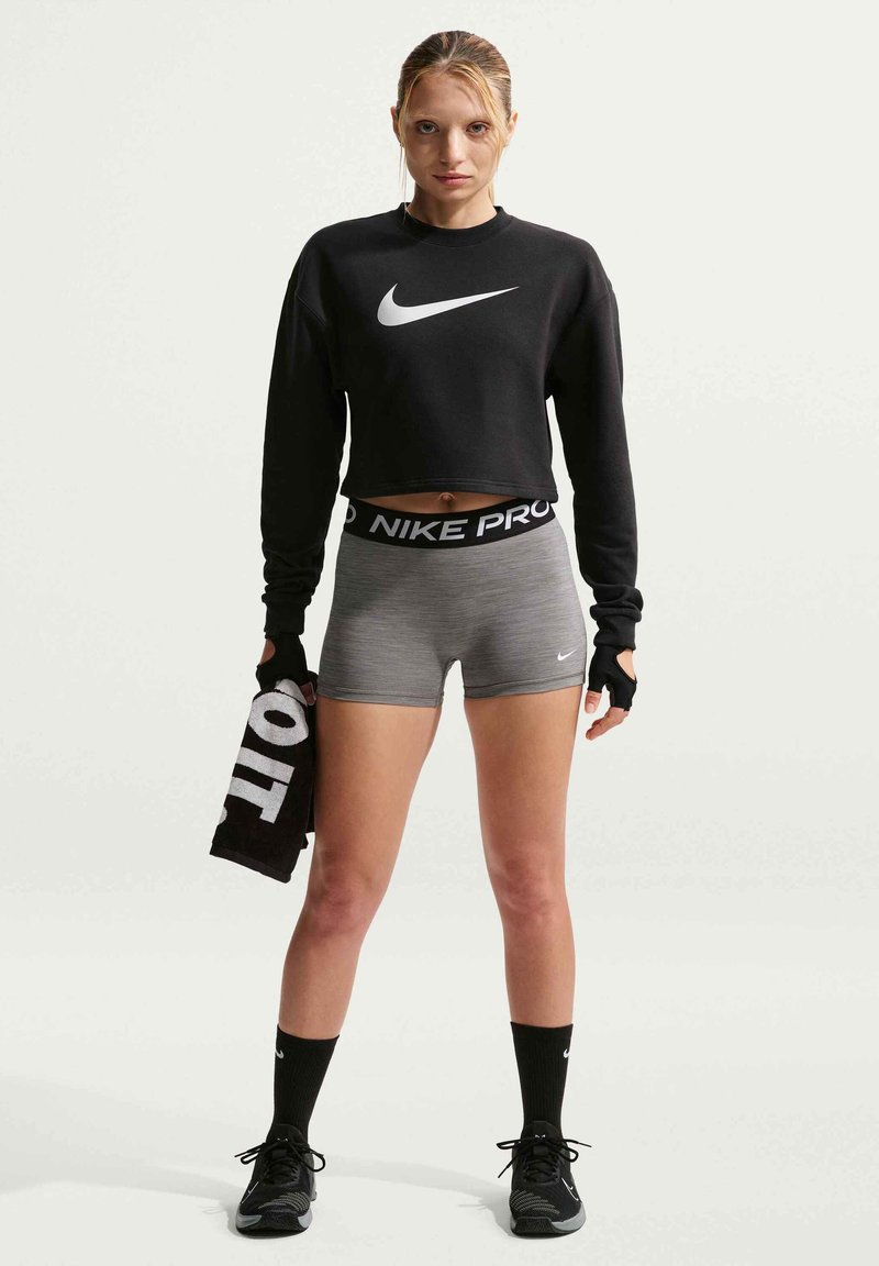 Frau in schwarzem, kurz geschnittenem Sweatshirt und grauen Nike Pro Shorts hält ein schwarzes Handtuch, trägt schwarze Handschuhe, Socken und Laufschuhe und steht.