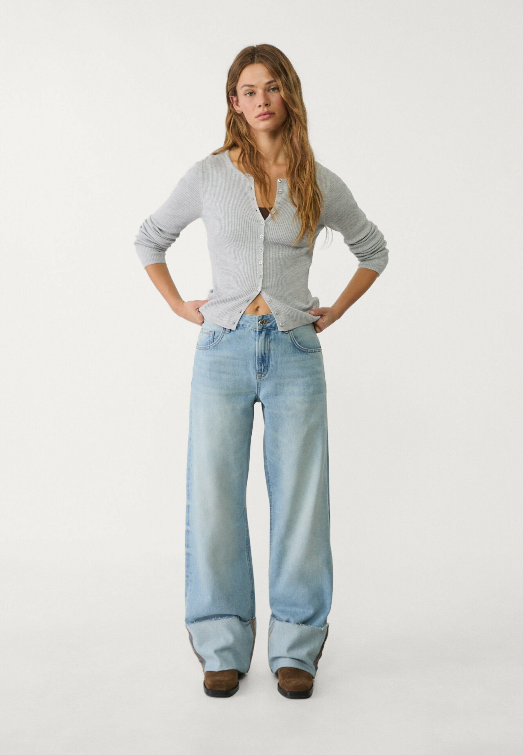 Hellgrauer, gerippter Cardigan mit Frontknöpfen, kombiniert mit lockeren hellblauen Jeans und braunen Ankle Boots. Sauberer weißer Hintergrund.