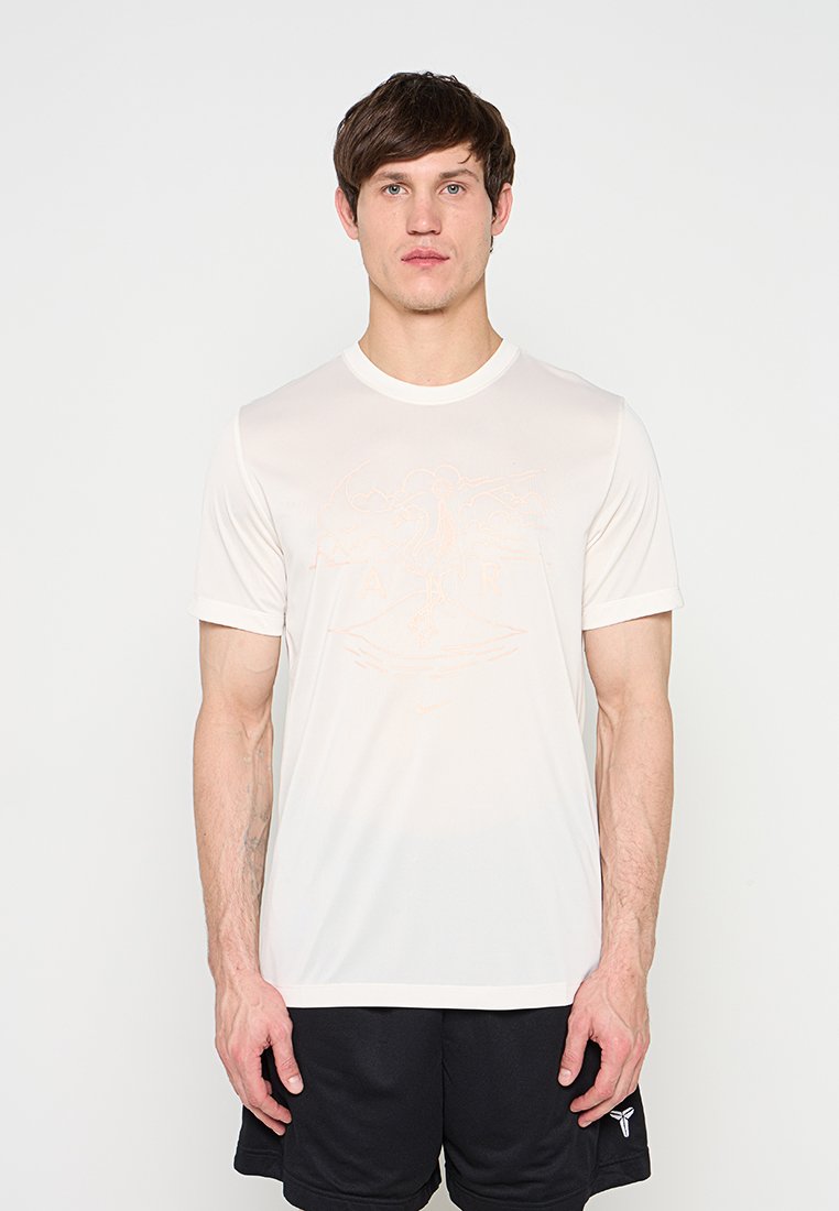 Mann trägt ein hellbeiges T-Shirt mit Grafik und schwarze Shorts, steht vor einem schlichten weißen Hintergrund, blickt nach vorne und hat eine entspannte Haltung.
