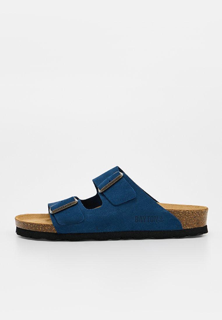 Blaue Wildleder-Slidesandale mit Korksohle, zwei verstellbaren Metall-Schnallenriemen und schwarzer Gummiaußensohle sichtbar vor schlichten Hintergrund.