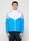 Mann steht frontal, trägt eine blau-weiße und graue Kapuzenjacke mit Reißverschluss und schwarze Nike-Hose vor einem grauen Hintergrund.