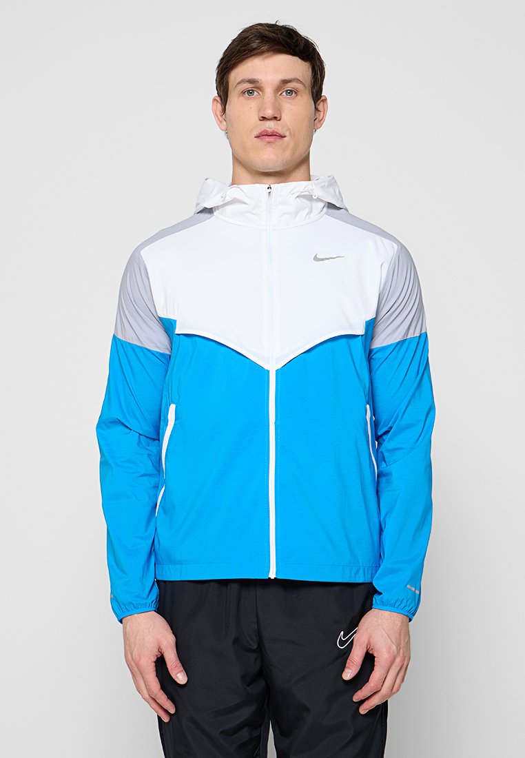 Mann steht frontal, trägt eine blau-weiße und graue Kapuzenjacke mit Reißverschluss und schwarze Nike-Hose vor einem grauen Hintergrund.
