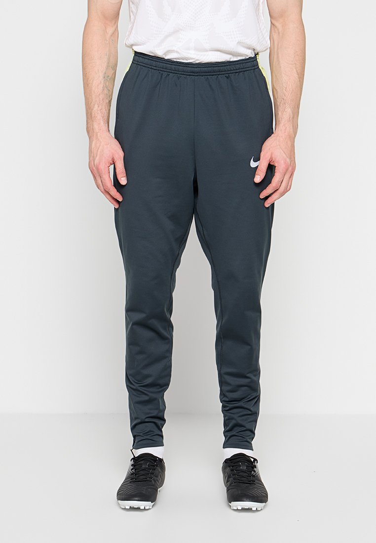Mann trägt dunkelgraue Nike-Sporthose, weiße Socken, schwarze Spikes, steht vor einem schlichten weißen Hintergrund.