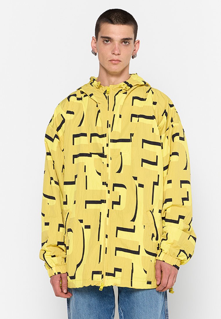 Gelbe Regenjacke mit großen schwarzen geometrischen Mustern, einem Frontreißverschluss und einer verstellbaren Kapuze. Der Stoff wirkt leicht und strukturiert.